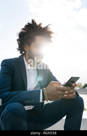 Young businessman sitting outdoors in sunshine using cell phone Stock Photo