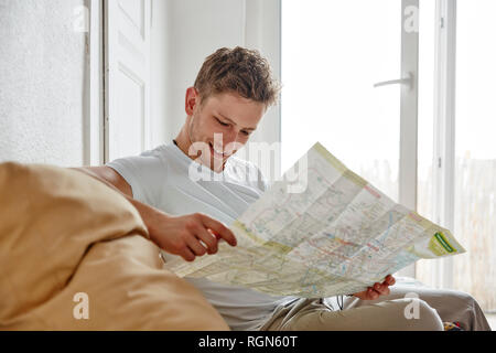 Smiling young man sitting on couch at home reading map Stock Photo