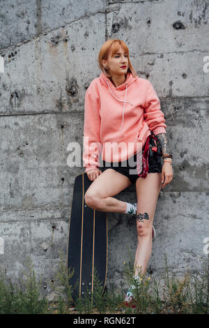 Portrait of cool young woman with carver skateboard leaning against a concrete wall Stock Photo