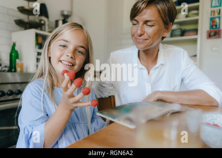 Happy mother and daughter sitting at table in kitchen at home playing with raspberries Stock Photo