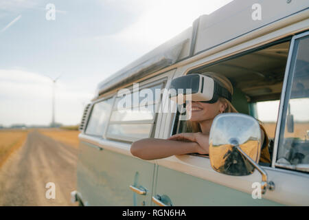 Smiling woman wearing VR glasses leaning out of window of a camper van Stock Photo