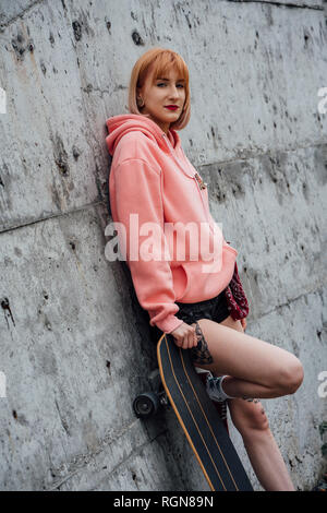 Portrait of cool young woman with carver skateboard leaning against a concrete wall Stock Photo