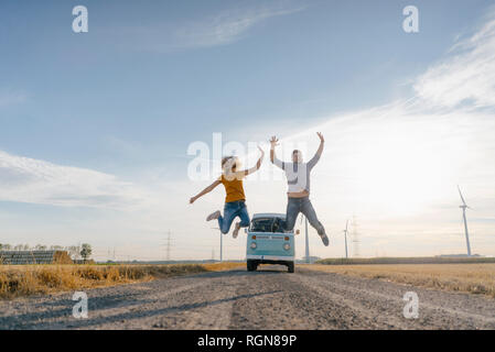 Exuberant couple jumping on dirt track at camper van in rural landscape