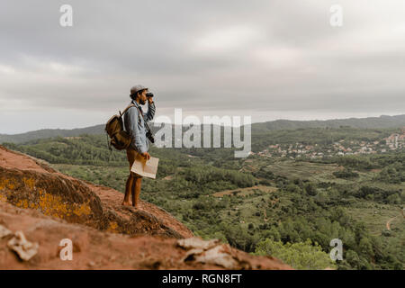 Young man with backpack, standing on a mountain, looking at view, using field glasses Stock Photo