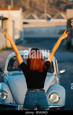 Redheaded woman standing infront of vintage car making victory sign, rear view Stock Photo