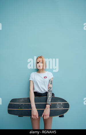 Portrait of cool young woman holding carver skateboard standing at turquoise wall Stock Photo