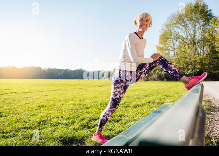 Smiling senior woman stretching on a bench in rural landscape