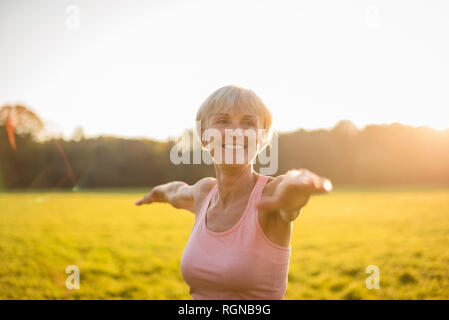 Portrait of smiling senior woman doing yoga on rural meadow at sunset Stock Photo