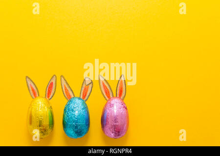 Three chocolate eggs in colorful aluminium foil with painted easter bunny ears on yellow background Stock Photo