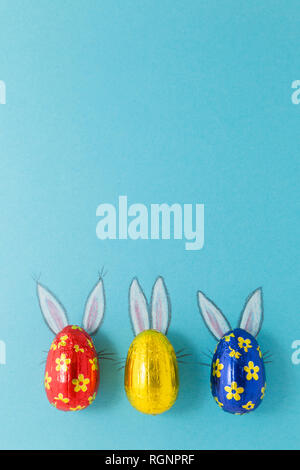 Three chocolate eggs in colorful aluminium foil with painted easter bunny ears on blue background Stock Photo