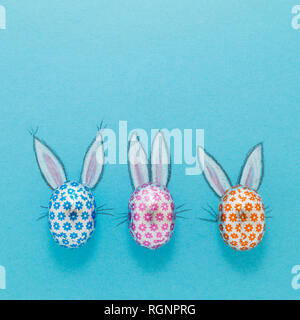 Three chocolate eggs in colorful aluminium foil with painted easter bunny ears on blue background Stock Photo
