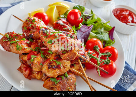 delicious chicken kebabs chargrilled on skewers served with herbs salad, tomatoes, lemon wedges on a white plate, on a wooden table, view from above