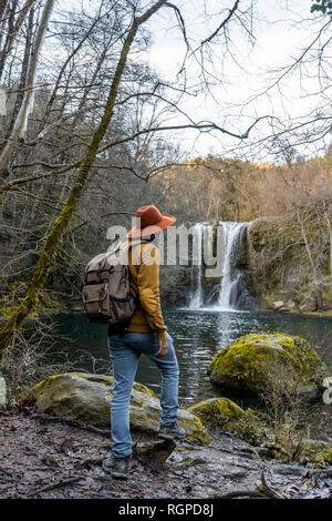 Back view of man with backpack stretching out arms and admiring beautiful waterfall while standing on boulder near forest lake in Spain Stock Photo