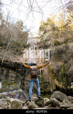 Back view of male traveler with outstretched arms standing on stones and admiring fantastic waterfall in forest in Spain Stock Photo