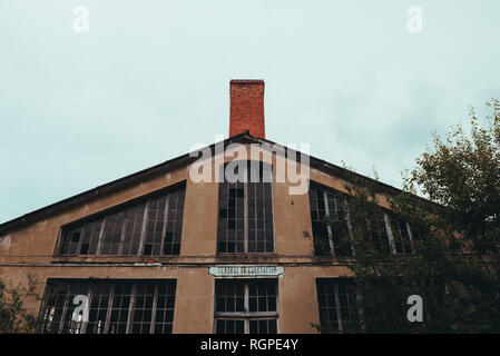 From below red facade of aged broken construction with big windows and chimney near trees and cloudy heaven Stock Photo