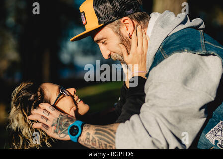 Side view of bearded man holding head of cheerful young lady in eyeglasses in park on blurred background Stock Photo