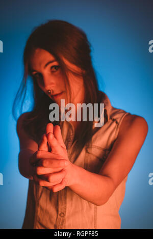 Charming young lady with piercing showing gun gesture and looking at camera in redness on blue background Stock Photo