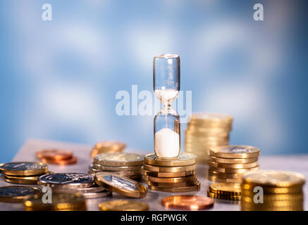 Sand trickles through an hourglass standing on a table with different coins. Stock Photo
