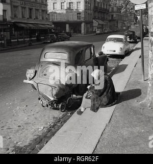 1950s, Paris, France, elderly lady, possibly a bag lady or vagabond, wearing a fur coat sitting beside a road and by parked cars with a small pram and her possessions with whilst reading a book. Stock Photo