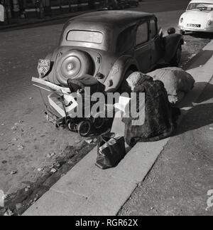 1950s, Paris, France, elderly lady, possibly a bag lady or vagabond, wearing a fur coat sitting beside a road and by parked cars with a small pram and her possessions with whilst reading a book. Stock Photo