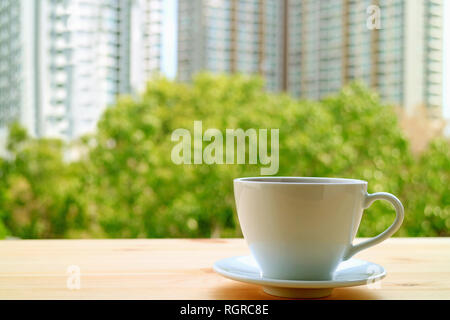 A cup of hot drinks on the window side wooden table with blurry green foliage and high buildings in background Stock Photo