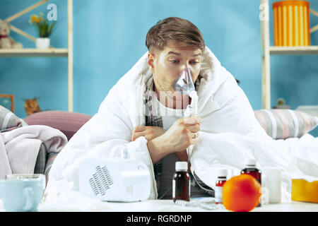 Bearded sick man with flue sitting on sofa at home covered with warm blanket and using an inhaler when coughing. The illness, influenza, pain concept. Relaxation at Home. Healthcare Concepts. Stock Photo