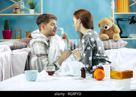 Bearded sick man and his wife sitting on sofa at home covered with warm blanket and using an inhaler when coughing. The illness, influenza, pain concept. Relaxation at Home. Healthcare Concepts. Stock Photo