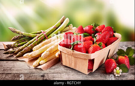 Wooden punnet of strawberries with fresh white and green asparagus shoots tied in bundles on an outdoor table in the garden or at farmers market