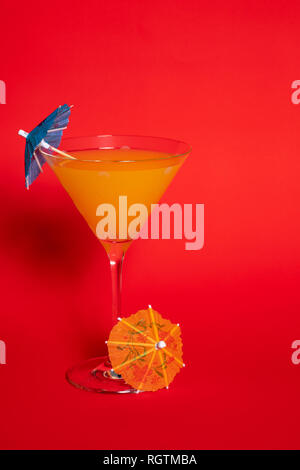 Orange drink with a blue umbrella in a martini glass set against a solid red background. An orange umbrella lies at the base of the glass. Stock Photo