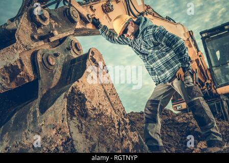 Construction Industry. Heavy Equipment Operator Job. Excavator and Caucasian Worker in Hard Hat. Men Looking Inside Excavator Bucket. Stock Photo