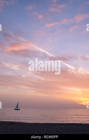 Small sailboat on gentled rippled sea sails into a delicate orange, pink and yellow sunset Stock Photo