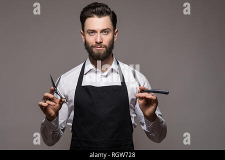 Portrait of cheerful, joyful barber with stubble in shirt having scissors, tools, equipments in hands looking at camera on grey background Stock Photo