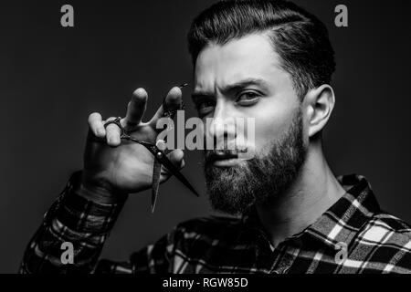 Young handsome bearded man with long beard moustache and brunette hair holding hairdresser or barber scissors with emotional face in studio on grey ba