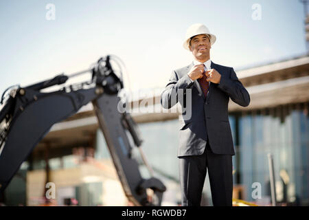 Young engineer wearing a suit and hardhat adjusts his tie while standing in front of a digger at a construction site. Stock Photo