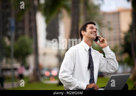 Mid-adult businessman laughing while using a laptop and talking on his cell phone. Stock Photo