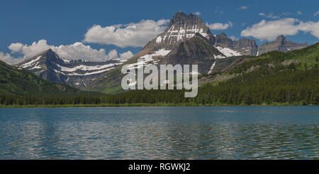 Mount Wilbur towers over surrounding mountains and the forested shore of Swiftcurrent Lake on a beautiful summer day in the Rocky Mountains in Glacier Stock Photo