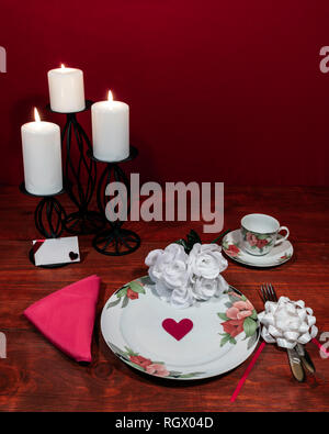 Floral pattern fine china dinnerware with matching plate, cup and saucer. bouquet of white roses, pink napkin, silverware, white candles and card Stock Photo