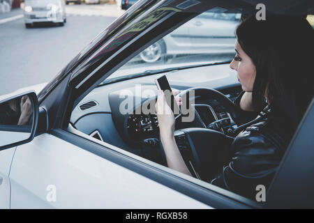 Woman using navigation app on smartphone while driving a car. Side view of brunette girl sitting in vehicle and looking at blank screen of cellphone.  Stock Photo