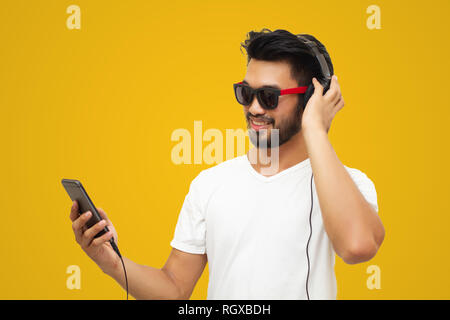 Asian handsome man with a mustache, smiling and laughing and using smart phone to listen music with headphones  on yellow backgound Stock Photo