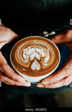 A cup of coffee with a heart latte foam art on top, being helf by masculine hands, warm brown colors, earth tones. Stock Photo