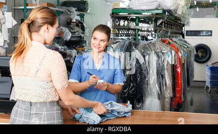 Young diligent positive woman worker of laundry taking clothes for dry cleaning from female client Stock Photo