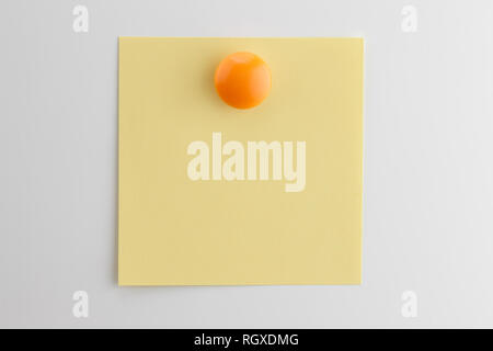 Square blank sheet of yellow paper pinned with orange button magnet on white refrigerator Stock Photo