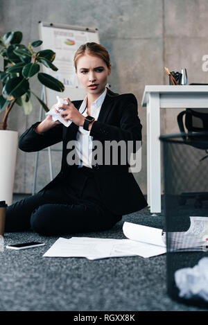 businesswoman sitting on floor and throwing crumpled papers to basket Stock Photo