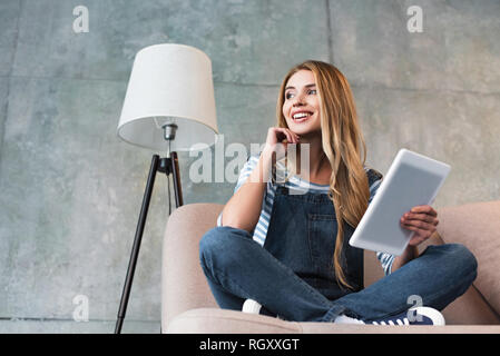 young smiling woman sitting on pink sofa in room with digital tablet in hand Stock Photo