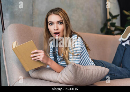 shocked young woman holding book in hands and lying on pink sofa Stock Photo