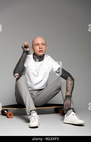 bald tattooed man sitting on skateboard and holding baseball bat on grey Stock Photo