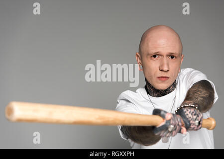 serious tattooed man hitting with baseball bat and looking at camera isolated on grey Stock Photo