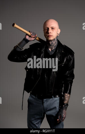 bald tattooed man in leather jacket holding baseball bat and looking at camera isolated on grey Stock Photo