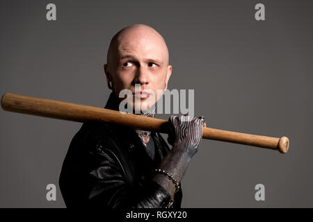 bald tattooed man in leather jacket holding baseball bat and looking away isolated on grey Stock Photo