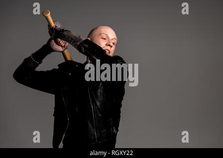 aggressive bald tattooed man in leather jacket hitting with baseball bat isolated on grey Stock Photo